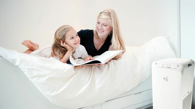 Hvit luftrenser fra Daikin plassert ved seng der mor og barn ligger og leser.